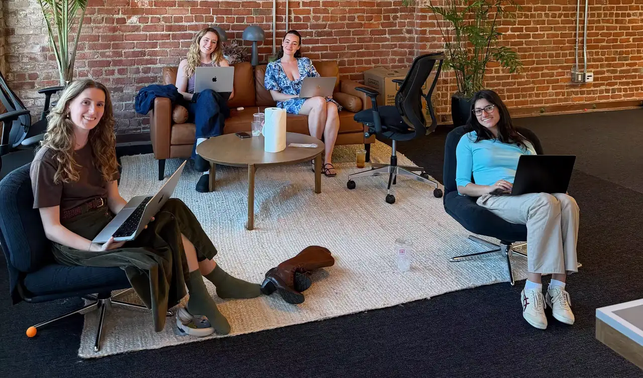Four teammates working on laptops from a leather sofa and armchairs in the San Francisco office, exposed brick behind them.