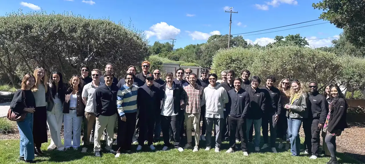 About thirty Tavrn teammates standing together on a lawn between olive trees at a team offsite.