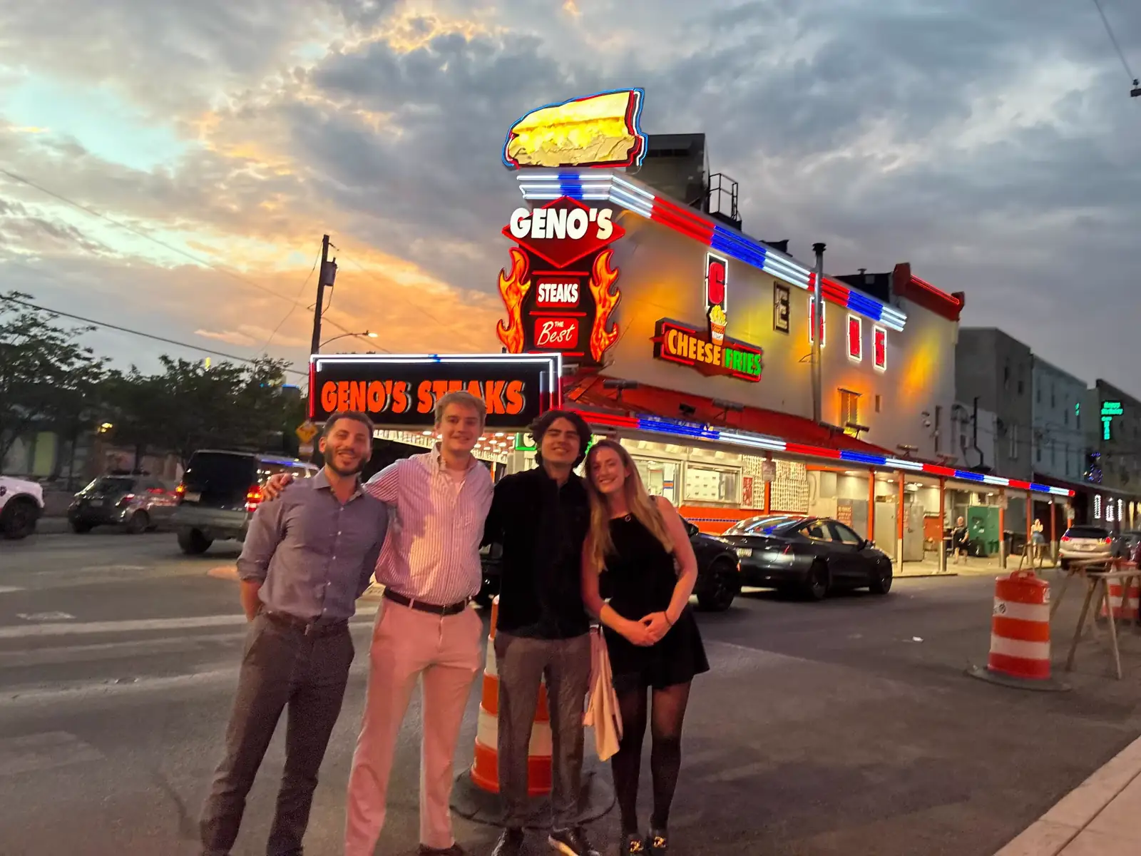Four teammates arm in arm on the sidewalk in front of Geno's Steaks in Philadelphia at dusk.